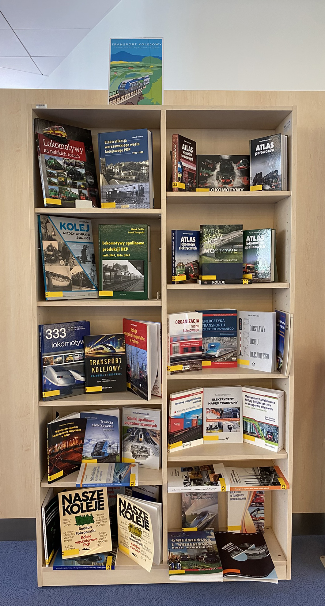  Zdjęcie przedstawia drewniany regał z książkami o tematyce kolejowej. Na półkach ułożone są liczne publikacje, w tym atlasy lokomotyw, książki historyczne i techniczne dotyczące transportu kolejowego oraz czasopisma branżowe. U góry regału wisi plakat z ilustracją pociągu, również o tematyce kolejowej.